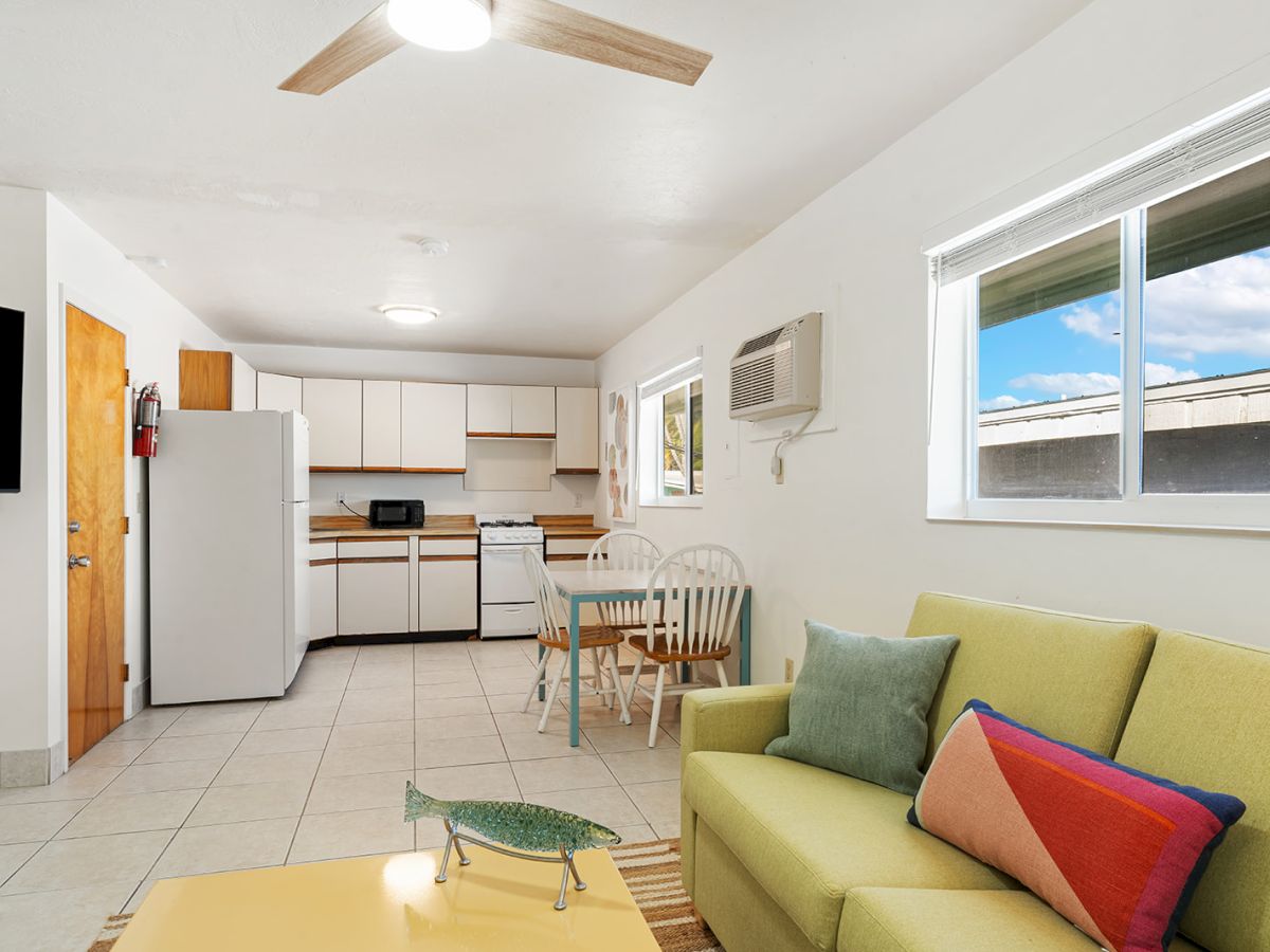 A bright, open-concept living area with a lime-green sofa, colorful pillows, and a glass coffee table, adjacent to a compact kitchen and dining space.