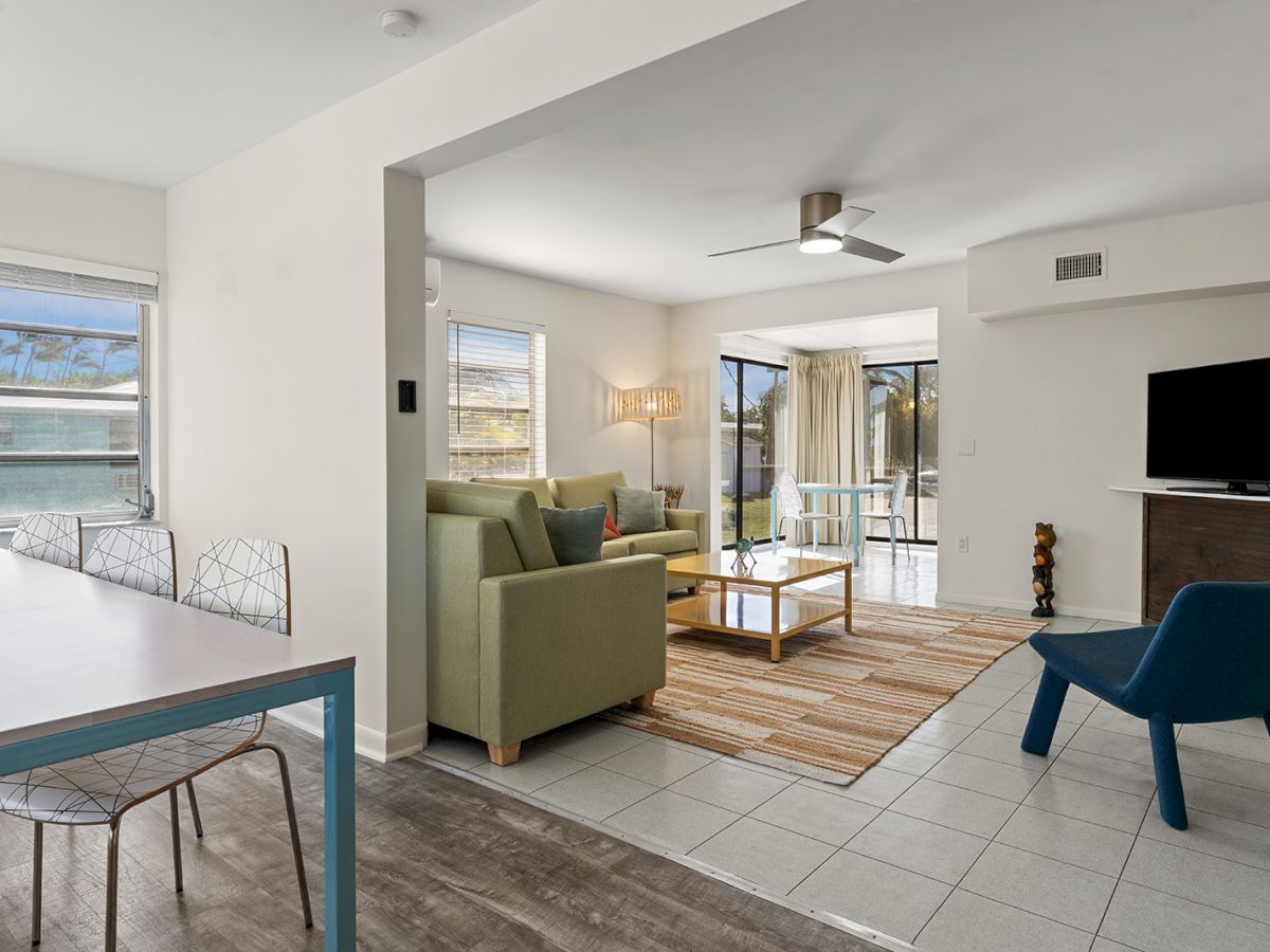 A bright, modern living area with a green sofa, blue chair, rug, wall-mounted TV, ceiling fan, and an open dining space with large windows.
